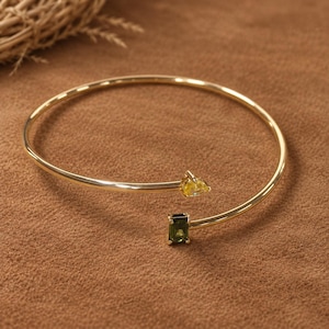 May include: A gold-toned open cuff bracelet featuring two distinct gemstones at the ends. One end holds a pear-shaped yellow stone, while the other end features a rectangular, emerald-cut green stone. The band is thin, polished, and circular.