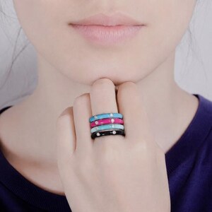 May include: A stack of four thin metal rings featuring smooth enamel finishes in bright blue, hot pink, light blue, and black. Each individual ring is accented with a single small, clear, round-cut gemstone set into the center of the band.