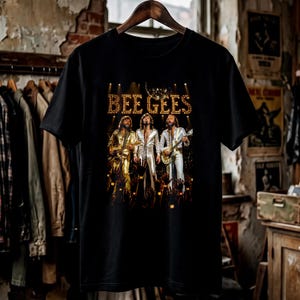 May include: A black short-sleeve t-shirt featuring a graphic print of the musical group Bee Gees performing on stage. The image shows three individuals in metallic outfits playing guitars and singing under stage lights with the text BEE GEES.