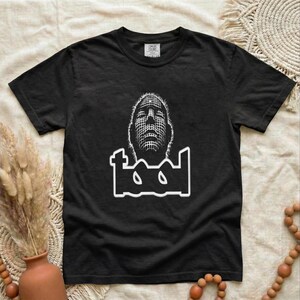 May include: A black short-sleeve t-shirt featuring a white graphic print of a stylized human face composed of a grid pattern, positioned above the word "tool" in a bold, custom white font. The shirt has a crew neckline and a standard fit.