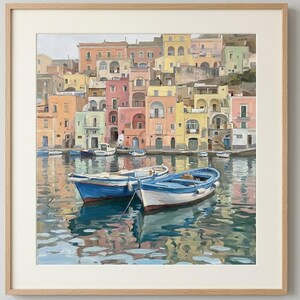 May include: A framed painting depicting a coastal scene with two small blue and white boats floating on calm water. In the background, a cluster of multi-coloured buildings in shades of yellow, pink, and peach rises along the waterfront.