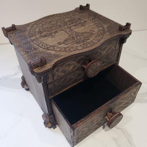 Può includere: Un forziere in legno marrone scuro con due cassetti, caratterizzato da intricati motivi a intreccio celtico incisi al laser e un disegno dell'albero della vita sulla parte superiore. Ogni cassetto ha un pomello circolare in legno e l'interno è foderato.