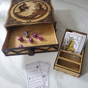 May include: A wooden box with a laser-engraved wizard and tower on the lid, featuring an open drawer containing six purple polyhedral dice. Beside it sits a wooden card holder with a deck of tarot cards and two loose instruction cards.