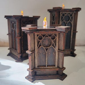 Pode incluir: Três castiçais de madeira castanho-escura com recortes arquitetónicos intrincados de estilo gótico, arcos apontados e motivos circulares. Cada estrutura retangular é rematada com uma pequena luz LED brilhante em forma de chama.