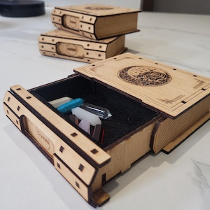 Peut inclure: Une boîte en bois clair en forme de livre avec un tiroir coulissant contenant plusieurs petits cristaux. La façade du tiroir porte l'inscription Crystals. Deux autres boîtes en bois en forme de livre sont empilées à l'arrière-plan.