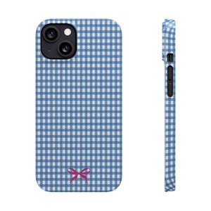Pode incluir: Uma capa de telefone com padrão xadrez vichy azul e branco com um laço rosa na parte inferior. A capa é mostrada na frente e na lateral, destacando o design e o recorte da câmera.