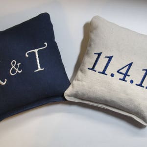 May include: Two square throw pillows. One is navy blue with white embroidered text reading K & T. The other is a natural beige color with navy blue embroidered text reading 11.4.17. Both pillows feature a simple, clean design with visible fabric texture.