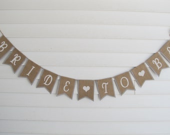 May include: A burlap banner with the words "Bride & Groom" in white lettering and two white hearts. The banner is strung on a white wall.