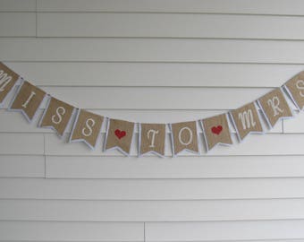 May include: A burlap banner with white letters that reads "Miss to Mrs" with red hearts between the words.