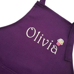 May include: A deep purple apron featuring the embroidered name Olivia in white script lettering, accompanied by a small, colorful cupcake graphic with pink, blue, and yellow details, including adjustable shoulder straps with black plastic sliders.
