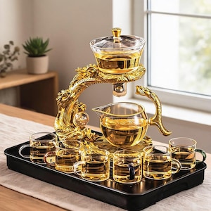 Puede incluir: Un juego de té dorado con forma de dragón que incluye una tetera de cristal y seis tazas pequeñas de cristal sobre una bandeja rectangular negra. La tetera está suspendida por una escultura de dragón metálica y el set incluye un infusor.