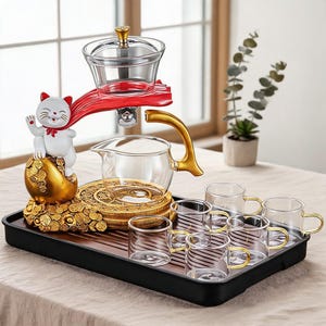 Puede incluir: Un juego de té de cristal que incluye una tetera giratoria con asa dorada, un infusor de cristal transparente y seis tazas pequeñas con asas doradas. Incluye una figura de gato blanco sobre monedas de oro en una bandeja negra.