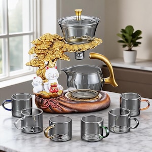 Puede incluir: Un juego de té con una tetera de cristal gris y seis tazas de cristal gris a juego con asas de colores. El conjunto incluye una base decorativa con forma de árbol dorado con dos figuras de gatos blancos y una bolsa roja de soporte.