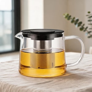 May include: A clear glass teapot with a cylindrical body, a black lid, and a matching black handle. Inside the teapot sits a stainless steel mesh infuser basket submerged in golden-colored tea. The teapot features a simple spout and a glass handle.