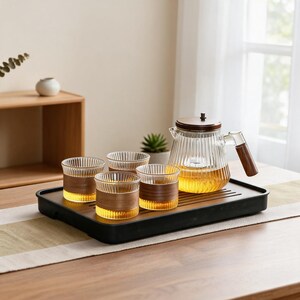 Puede incluir: Un juego de té de cristal que incluye una tetera acanalada con tapa y asa de madera oscura, junto con cuatro tazas de cristal acanalado a juego con fundas de madera oscura, dispuesto sobre una bandeja de servicio rectangular negra con té.