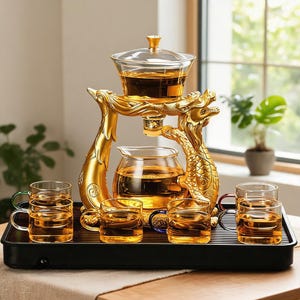Puede incluir: Un juego de té con una tetera e infusor de cristal transparente suspendidos por un soporte dorado con forma de dragón. El conjunto incluye seis tazas pequeñas de cristal con asas de colores, dispuestas sobre una bandeja rectangular negra.