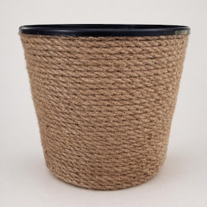 Può includere: Un vaso cilindrico con bordo nero ed esterno in corda intrecciata e strutturata. La corda marrone naturale è avvolta strettamente attorno al vaso, creando un aspetto rustico e decorativo. Adatto per uso interno o esterno.