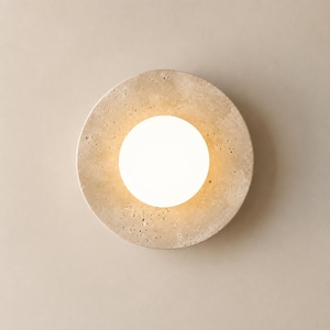May include: A circular wall light fixture with a central, bright white globe. The outer ring is a textured, beige stone. The light emits a warm glow. The fixture is designed to be mounted on a wall, providing ambient lighting.