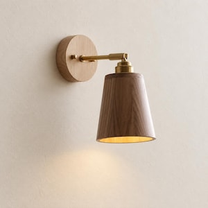 Peut inclure: Une applique murale composée d'une base circulaire en bois clair, d'un bras en métal couleur laiton et d'un abat-jour conique en bois foncé. Le luminaire émet une lueur chaude par le bas de l'abat-jour, soulignant la texture du grain du bois.