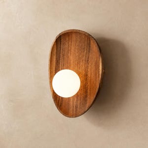 May include: A wall-mounted light fixture featuring a smooth, oval-shaped wooden backplate with a natural grain pattern. A single, spherical white glass globe bulb is positioned off-center on the surface of the wood, emitting a soft glow.