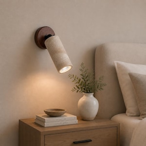 May include: A wall-mounted spotlight featuring a cylindrical beige travertine stone shade attached to a dark wood circular base. The light fixture is angled downwards, casting a warm glow over a stack of two books and a small ceramic bowl.