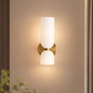 Peut inclure: Applique murale moderne avec une finition en laiton et deux abat-jours en verre blanc. L'applique a un abat-jour supérieur cylindrique et un abat-jour inférieur arrondi, reliés par un disque en laiton. Fixée sur un mur beige.