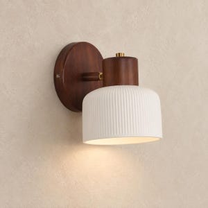 Peut inclure: Une applique murale composée d'une plaque de fixation circulaire en bois foncé et d'une pièce supérieure cylindrique assortie. Un abat-jour en céramique blanche à texture nervurée verticale est fixé, rehaussé d'un petit détail en métal doré.