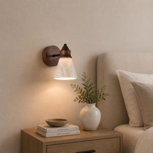 Puede incluir: Un aplique de pared con base circular de madera oscura, brazo de metal negro y pantalla cónica con acabado de mármol blanco. La luz emite un brillo cálido que ilumina el área inferior donde se encuentran unos libros y un florero.