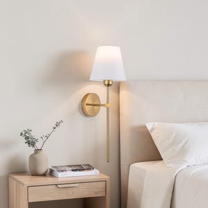 Peut inclure: Une applique murale dorée avec un abat-jour blanc est fixée sur un mur clair. L'applique a un long bras fin et une base circulaire. Un petit vase avec de la verdure et des livres sont sur une table de chevet en bois à côté d'un lit.