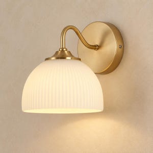 May include: A wall-mounted light fixture featuring a brushed brass circular base and a curved arm that supports a dome-shaped, white frosted glass shade with a vertical ribbed texture. The lamp emits a soft, warm glow from beneath the shade.