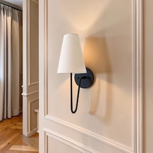 May include: A wall-mounted sconce featuring a white lampshade and a black metal arm and base. The sconce is fixed to a light beige wall, with a minimalist and modern design.
