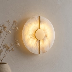 May include: A round, illuminated wall sconce with a marble-like appearance. The light fixture features a central, circular light source and is accented with vertical gold-colored metal strips. The sconce is mounted on a neutral-colored wall.
