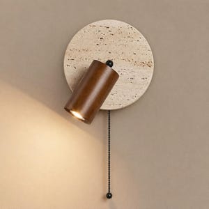 May include: A wall-mounted light fitting featuring a circular beige travertine stone backplate and a cylindrical dark brown wooden spotlight head. A black metal pull chain hangs vertically from the base of the wooden spotlight component.