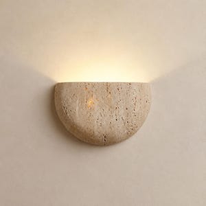 May include: A semi-circular wall sconce crafted from beige, porous travertine stone. The fixture features a textured, natural surface and emits a warm, upward-facing glow that illuminates the wall above it, creating a soft light effect.