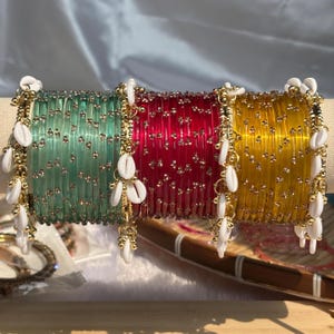 Puede incluir: Un conjunto de brazaletes translúcidos en verde menta, rojo intenso y amarillo dorado, cada uno con pequeños detalles metálicos en tono dorado. Los brazaletes están adornados con conchas cauri blancas colgantes en cadenas doradas.