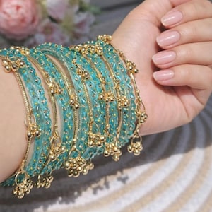 May include: A set of multiple translucent turquoise bangles featuring gold-toned metallic accents and small dangling gold-toned charms. The bracelets are arranged in a stack, showcasing a textured surface with embedded gold-colored flecks.