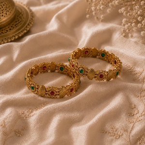 May include: Two gold-colored bangles with intricate designs and embedded red and green gemstones. The bangles are displayed on a cream-colored, textured fabric, with a decorative gold plate in the background.
