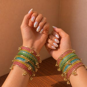 Peut inclure: Deux poignets ornés de bracelets colorés. Chaque ensemble de bracelets présente un éventail de bandes roses, vertes, bleues et jaunes, ornées de petites clochettes dorées. Les bracelets sont empilés, créant un effet de superposition. Le fond est de ton neutre et chaud.