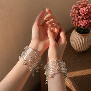 Op de afbeelding: Een set van meerdere doorzichtige, flexibele kunststof armbanden die om elke pols worden gedragen. Elke armband is voorzien van kleine, bungelende zilverkleurige metalen bedeltjes die met tussenpozen aan de ring zijn bevestigd.