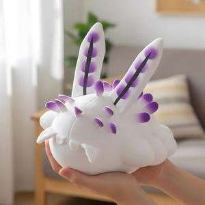 May include: A white plush toy shaped like a sea slug, featuring two prominent upright sensory rhinophores with purple tips and a dark central line, along with smaller purple-tipped protrusions along the rounded body and a soft, smooth texture.