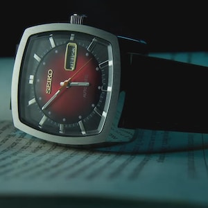 Puede incluir: Un reloj automático Seiko vintage con una caja plateada rectangular y una esfera burdeos. El reloj presenta una correa de cuero negro y está apoyado sobre un libro. La esfera muestra el día y la fecha.