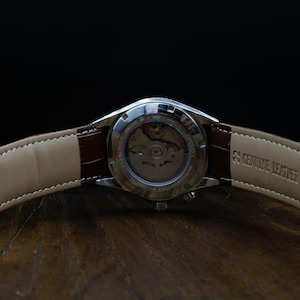 Pu&ograve; includere: Primo piano di un orologio da polso con cinturino in pelle marrone. L'orologio ha una cassa argentata e un retro trasparente che rivela il meccanismo. Il cinturino &egrave; contrassegnato con "Genuine Leather". L'orologio &egrave; su una superficie di legno.