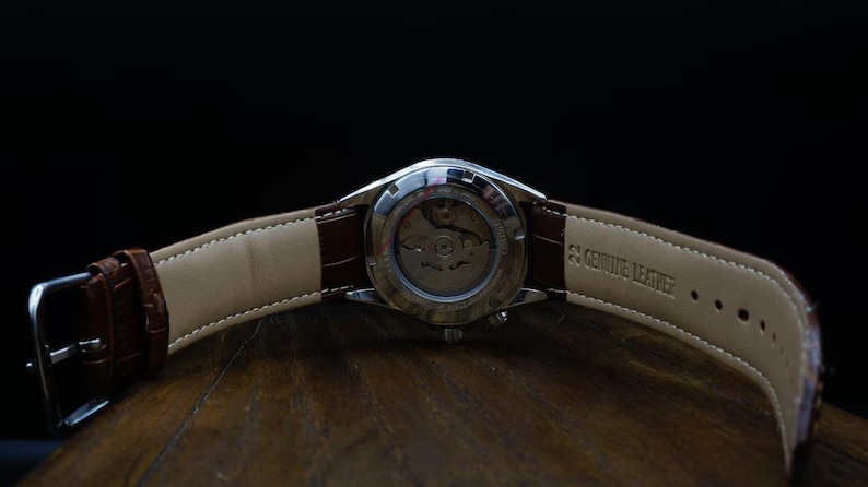 Pu&ograve; includere: Primo piano di un orologio da polso con cinturino in pelle marrone. L'orologio ha una cassa argentata e un retro trasparente che rivela il meccanismo. Il cinturino &egrave; contrassegnato con "Genuine Leather". L'orologio &egrave; su una superficie di legno.