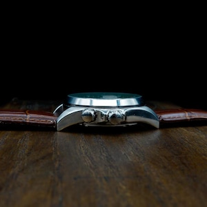 Pu&ograve; includere: Un orologio da polso argentato con cinturino in pelle marrone. Il quadrante dell'orologio &egrave; scuro e il cinturino presenta un motivo strutturato a forma di coccodrillo. L'orologio &egrave; posto su uno sfondo scuro su una superficie di legno.