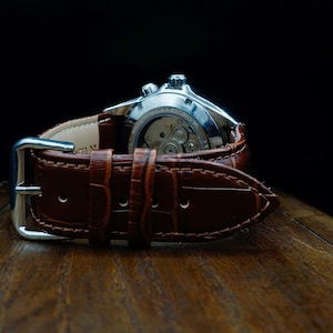Pu&ograve; includere: Primo piano di un orologio da polso con cinturino in pelle marrone. Il quadrante dell'orologio &egrave; argentato e il cinturino presenta un motivo strutturato, simile alla pelle di coccodrillo. L'orologio &egrave; su una superficie di legno.