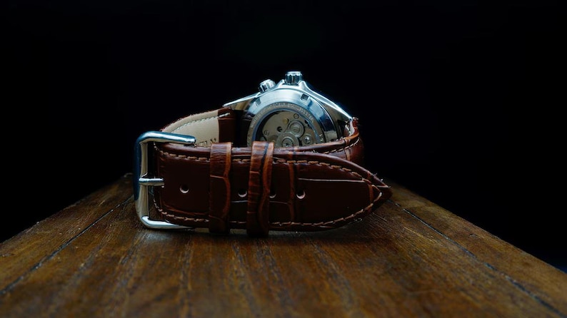 Pu&ograve; includere: Primo piano di un orologio da polso con cinturino in pelle marrone. Il quadrante dell'orologio &egrave; argentato e il cinturino presenta un motivo strutturato, simile alla pelle di coccodrillo. L'orologio &egrave; su una superficie di legno.