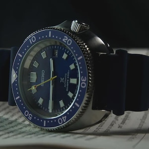 May include: A close-up of a Seiko automatic diver's watch with a blue dial and bezel. The watch has a black rubber strap and is set against a blurred background. The watch face displays the Seiko logo and other markings.