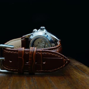 Pu&ograve; includere: Primo piano di un orologio da polso argentato con cinturino in pelle marrone. Il meccanismo interno dell'orologio &egrave; visibile. Il cinturino marrone ha una fibbia ed &egrave; su uno sfondo scuro.