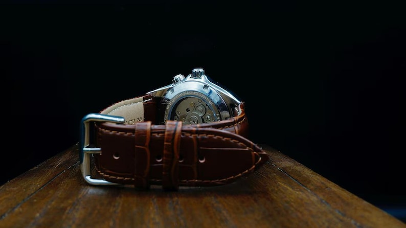 Pu&ograve; includere: Primo piano di un orologio da polso argentato con cinturino in pelle marrone. Il meccanismo interno dell'orologio &egrave; visibile. Il cinturino marrone ha una fibbia ed &egrave; su uno sfondo scuro.