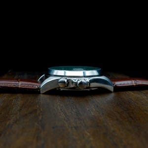 Pu&ograve; includere: Un orologio da polso argentato con cinturino in pelle marrone. Il quadrante dell'orologio &egrave; scuro e il cinturino presenta un motivo strutturato a forma di coccodrillo. L'orologio &egrave; posto su uno sfondo scuro su una superficie di legno.